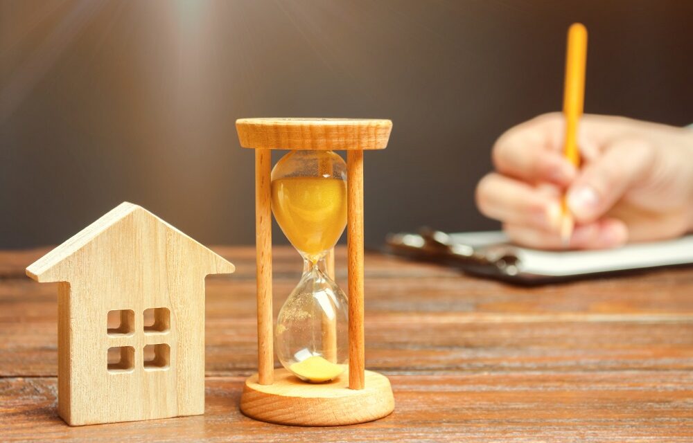 Wooden House And Clock. A Person Signs Documents. Signing A Contract To Rent A House Or Apartment. Making A Will. Testament Property. Buying Or Selling Real Estate. Mortgage Agreement. Leasing